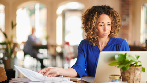 Holiday Temp Jobs Blog Graphic that showcases a woman in a blue button up sitting at a table with papers and her laptop.