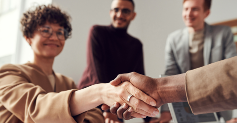 Photo of a woman smiling and shaking someone's hand. There are two people behind her.