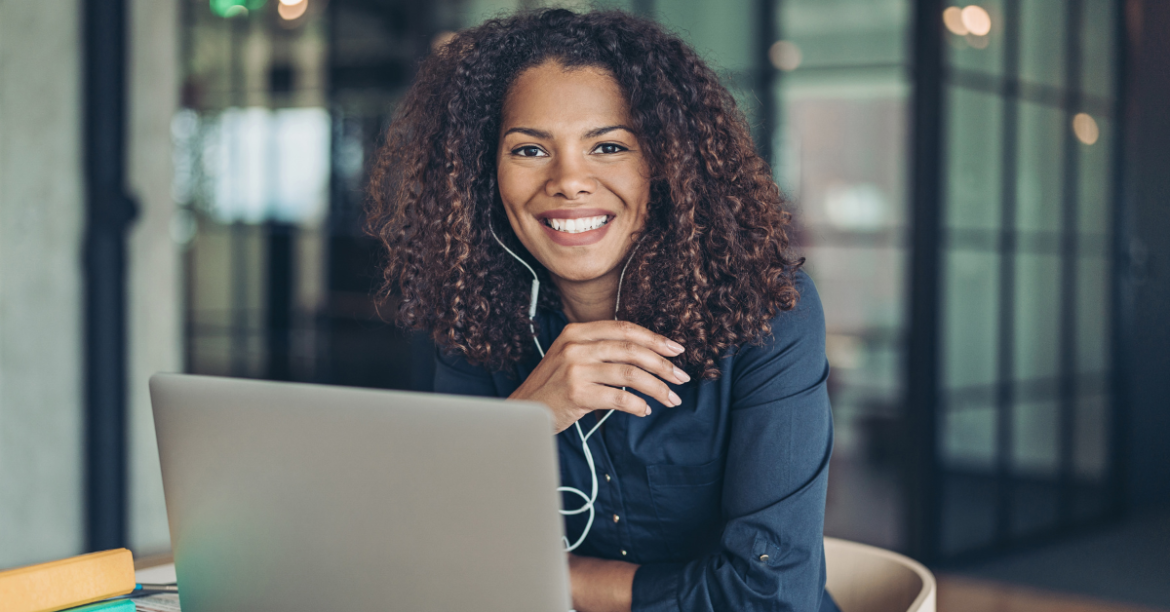 Black woman with curly hair sitting at a desk, smiling in front of her laptop